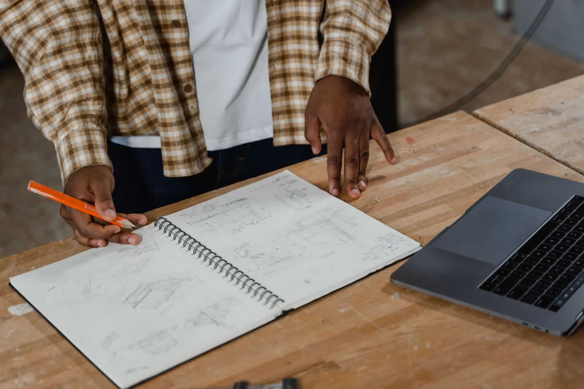 Hände skizzieren mit Bleistift in ein Notizbuch, daneben ein Laptop